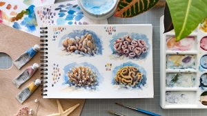 Ein Block mit Spiralbindung zeigt vier verschiedene Korallen gemalt mit Aquarell in bräunlichen und gelblichen Farben. Drum herum liegen verschiedene Kunstmaterialien wie Pinsel, Paletten und Farbtuben.